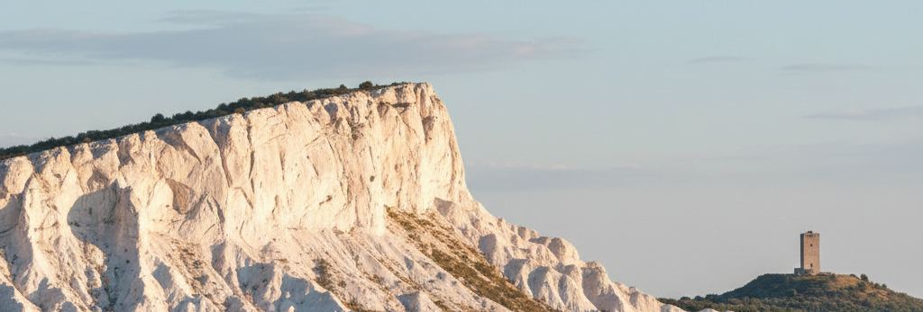 A white chalk cliff with low scrub along its rim, a square stone tower on a hill beyond, pale sky