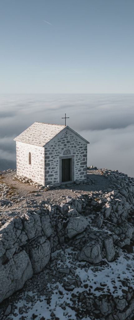 A small stone chapel with a metal cross on its roof, on a snow patched rock summit above a flat white overcast layer
