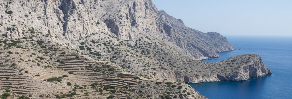 Bare limestone slopes cut with old stone terraces, falling to a rocky headland and deep blue sea under pale sky